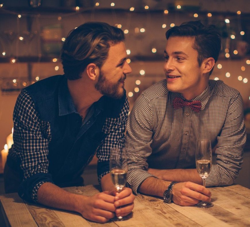 Loving gay couple having romantic date, drinking champagne. Domestic kitchen. Evening scene lit by candle and string lights.